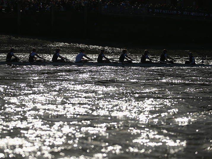 A crew rows across glimmering water on the Thames.